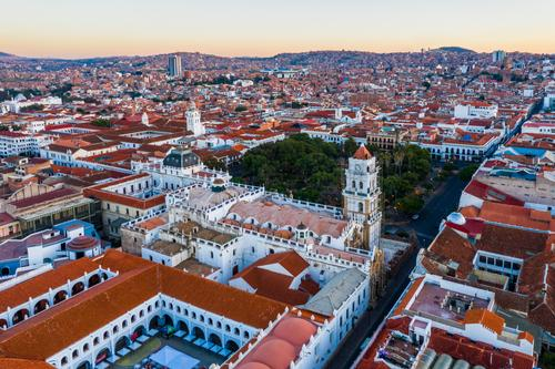 A panoramic aerial view of Sucre showing the cityscape, colonial buildings, and surrounding Andes mountains in Bolivia.