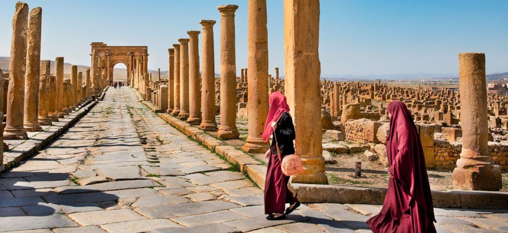 Visitors walking among the well-preserved Roman ruins at Timgad beneath a clear blue sky.