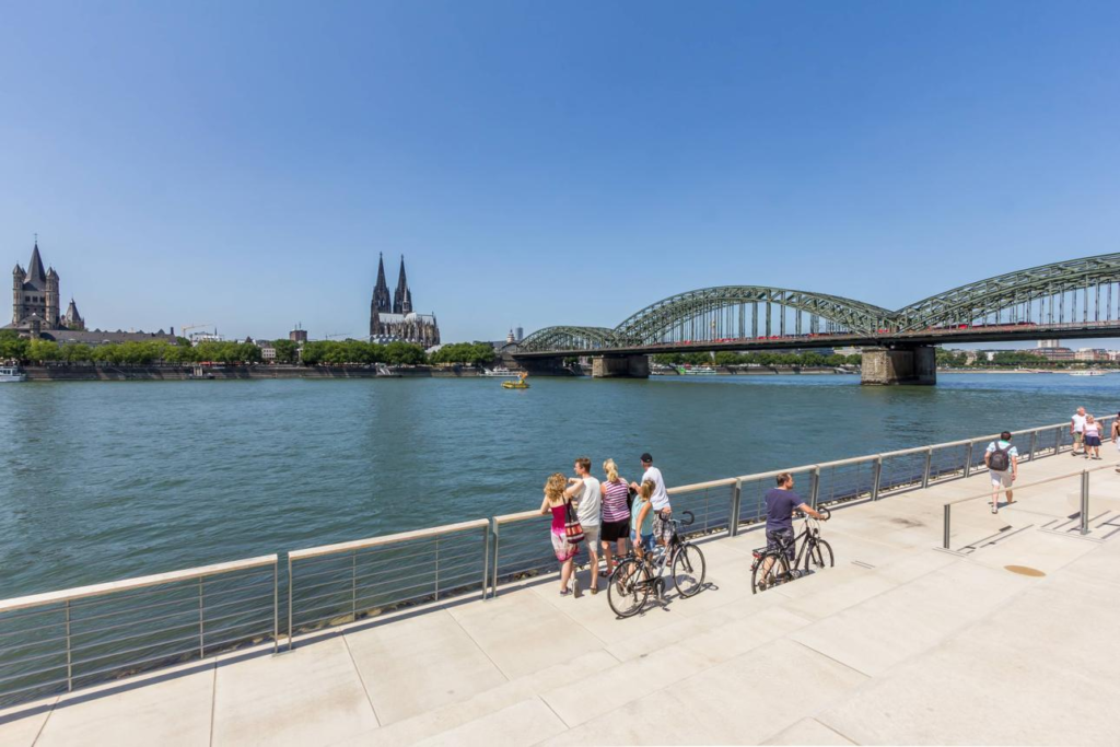 People enjoying a riverside promenade along the Rhine with city buildings in the background.