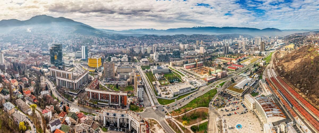 A panoramic view of Sarajevo with red-roofed buildings surrounded by forested mountains.