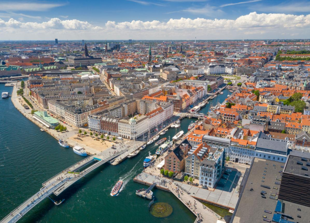 A panoramic view of Copenhagen featuring colorful waterfront buildings, historic rooftops, and boats along the harbor.